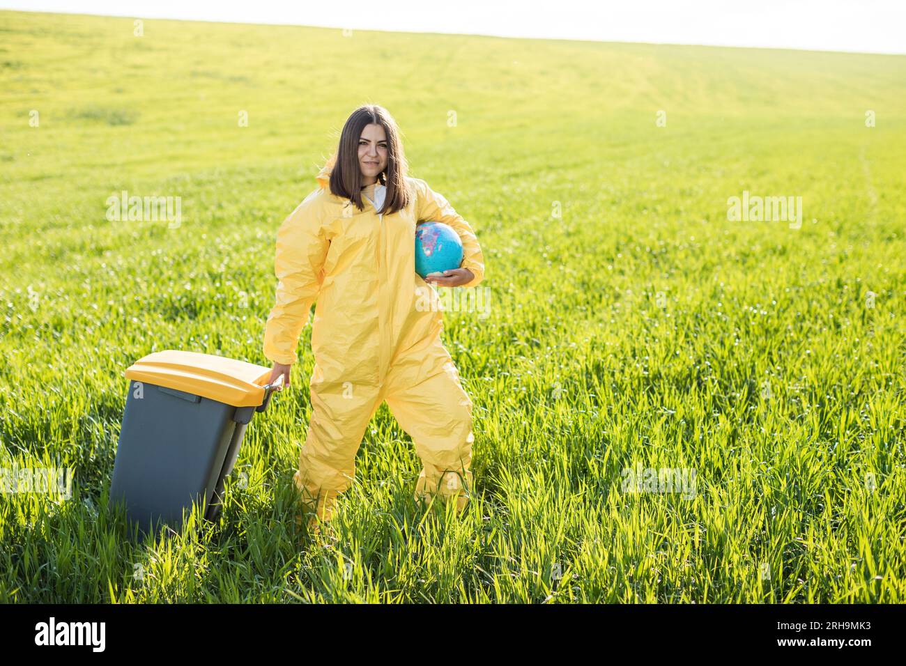 Una mujer en un traje protector amarillo está en medio de un campo verde sosteniendo un globo, y con su otra mano lleva una papelera. Foto de stock