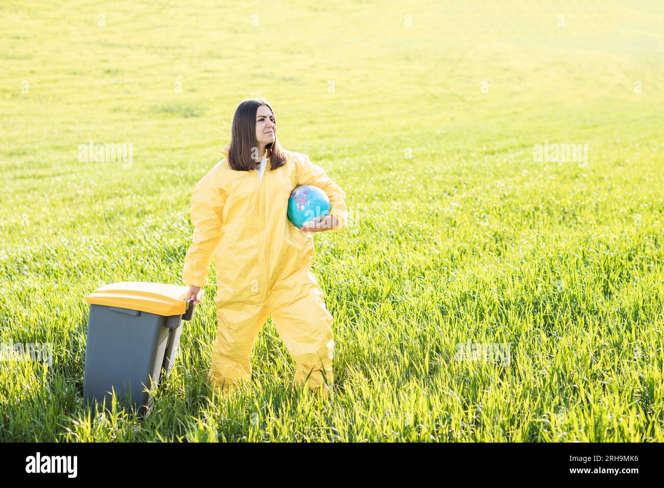 Una mujer en un traje protector amarillo está en medio de un campo verde sosteniendo un globo, y con su otra mano lleva una papelera. Foto de stock