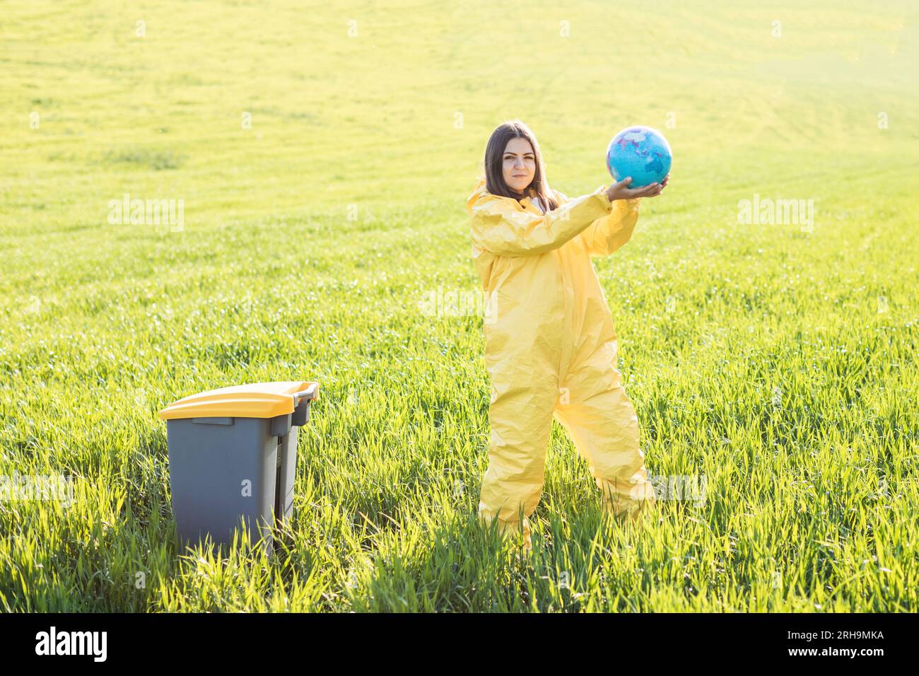 Una mujer en un traje protector amarillo está en medio de un campo verde y sostiene un globo en sus manos, junto a una papelera. El cuidado del planeta, excepto el Foto de stock