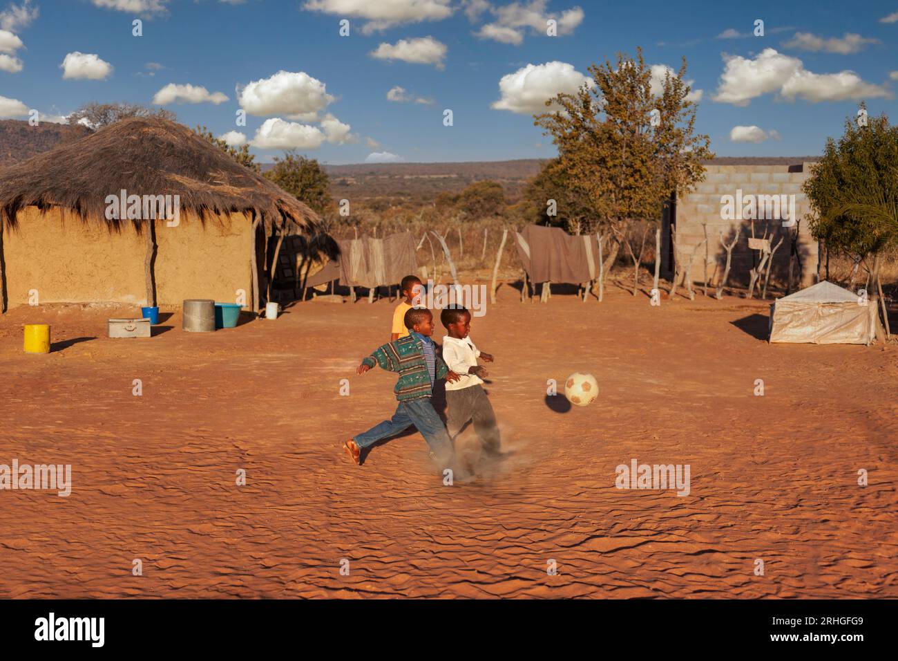 niños africanos de la aldea que juegan al fútbol en un campo de fútbol improvisado en el patio entre las casas de las cabañas de paja Foto de stock