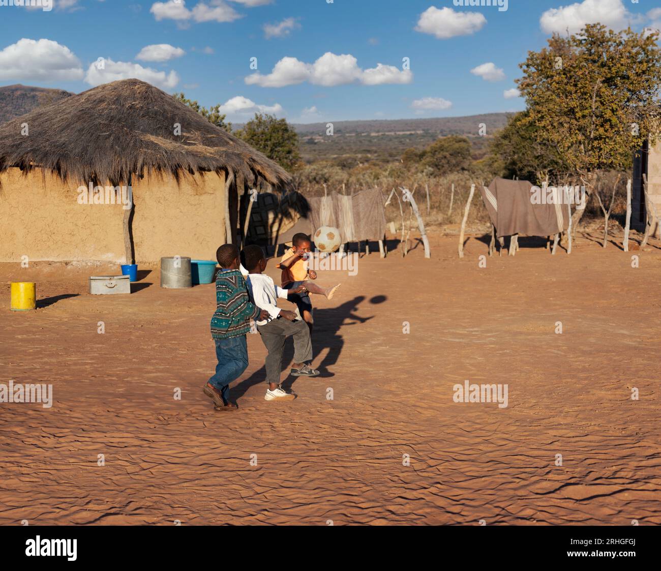 niños africanos de la aldea que juegan al fútbol en un campo de fútbol improvisado en el patio entre las casas de las cabañas de paja Foto de stock