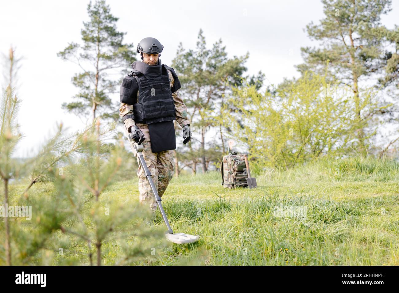Un hombre con uniforme militar y chaleco antibalas se sienta en el bosque  cerca de un detector de metales.