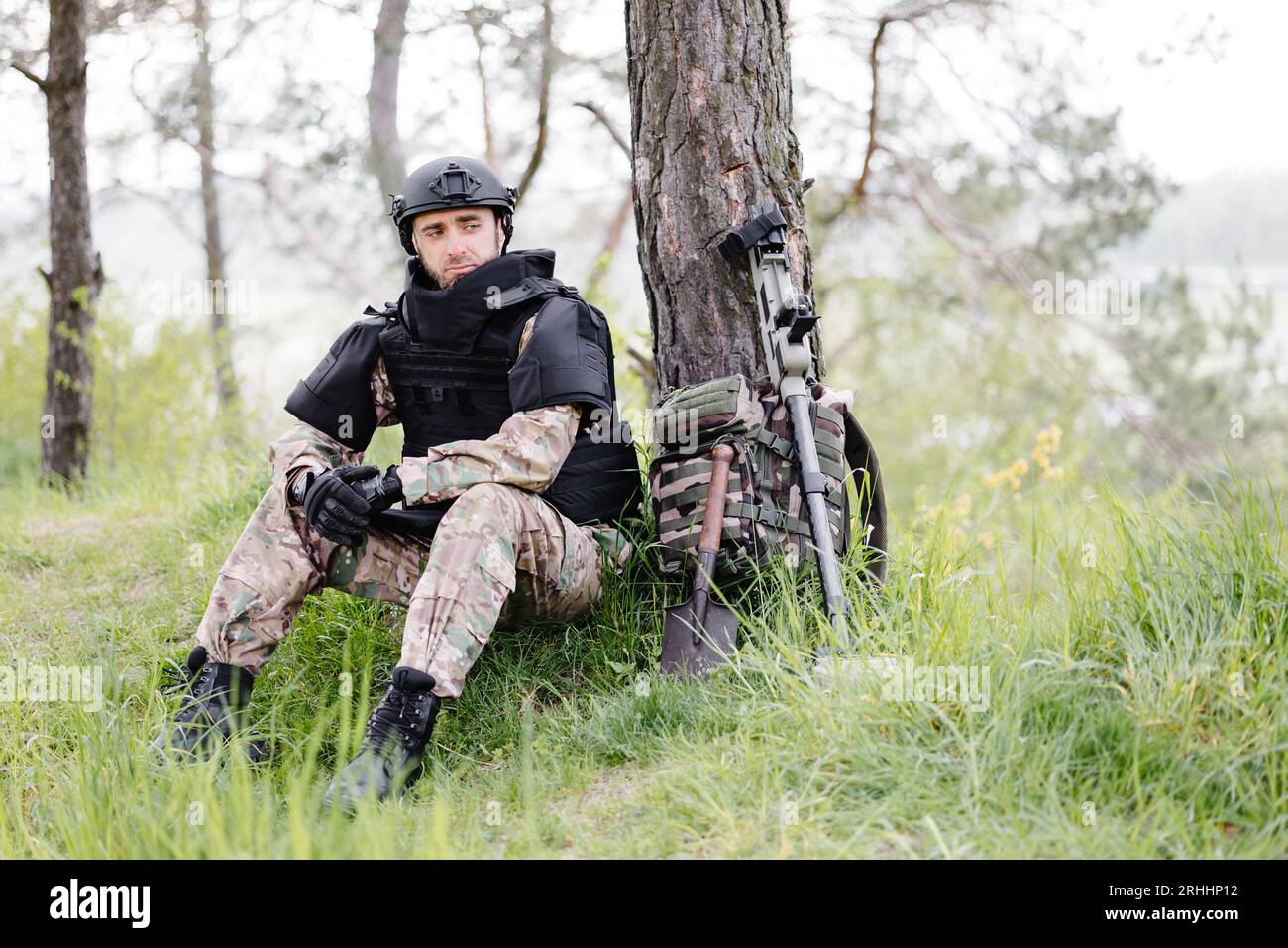 Un hombre con un uniforme militar y un chaleco antibalas se sienta en el  bosque cerca de un detector de metales y una mochila militar. Un hombre se  detiene en el trabajo