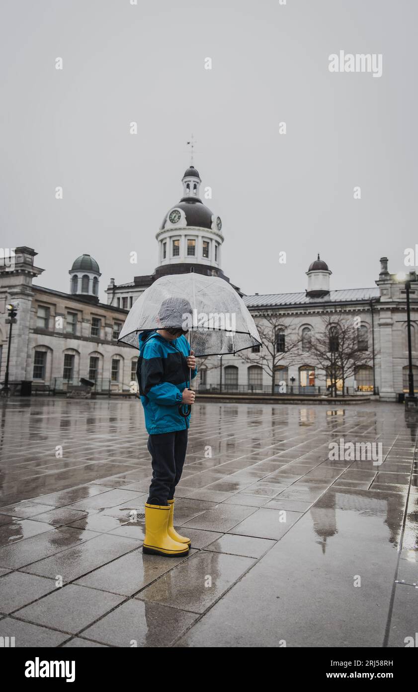 Muchacho en botas de lluvia amarillas sosteniendo paraguas en la lluvia en la plaza de la ciudad. Foto de stock