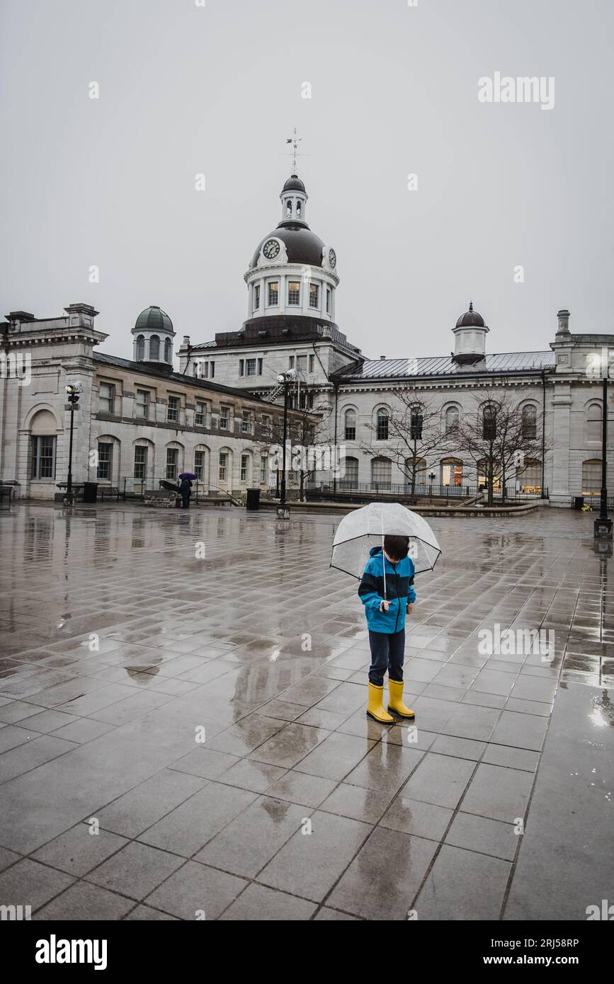 Muchacho en botas de lluvia amarillas sosteniendo paraguas en la lluvia en la plaza de la ciudad. Foto de stock