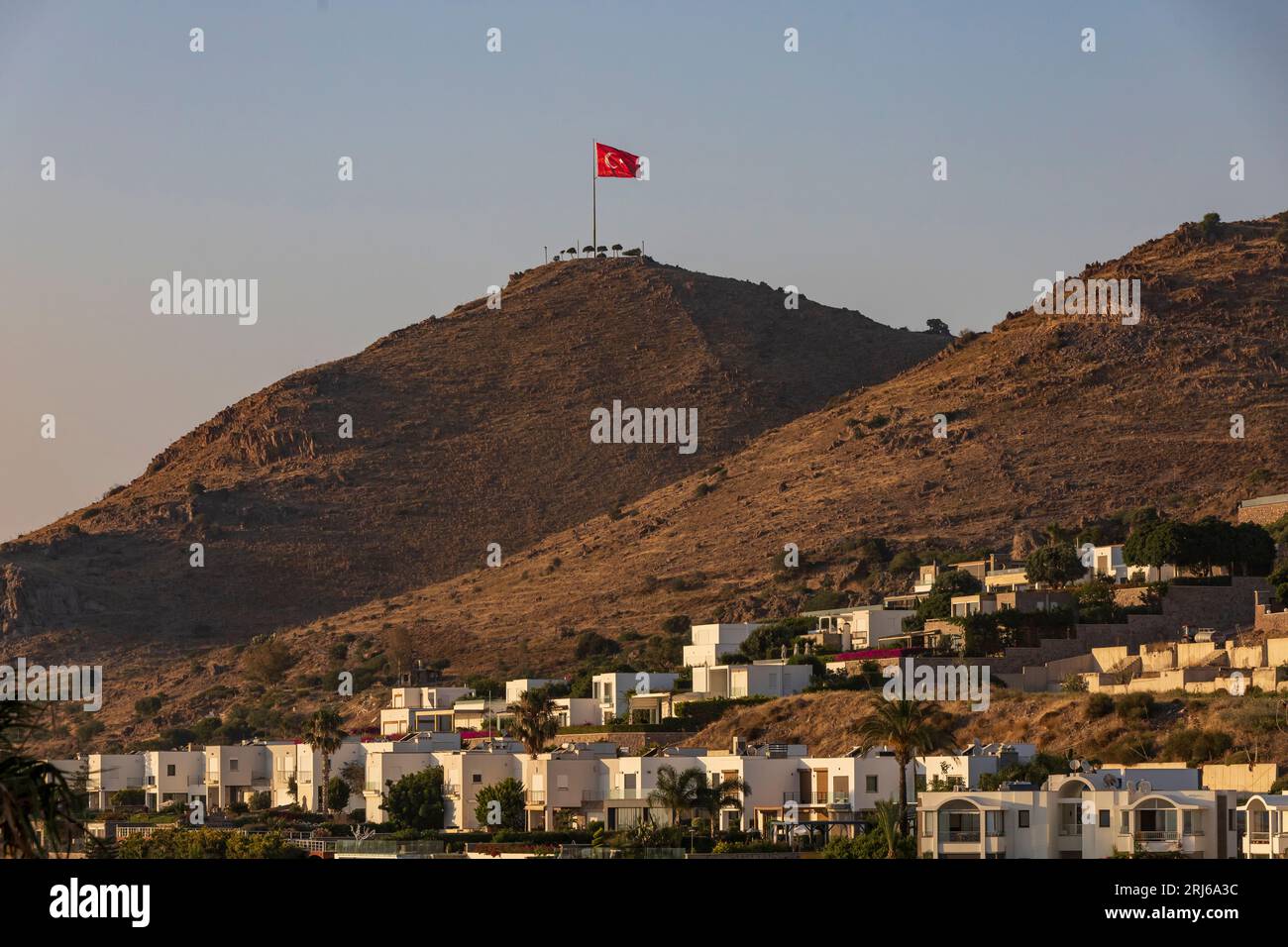 Bandera turca levantada en la cima de una colina con casas de verano unifamiliares en las laderas de Turgutreis, un centro de verano del Egeo. Foto de stock