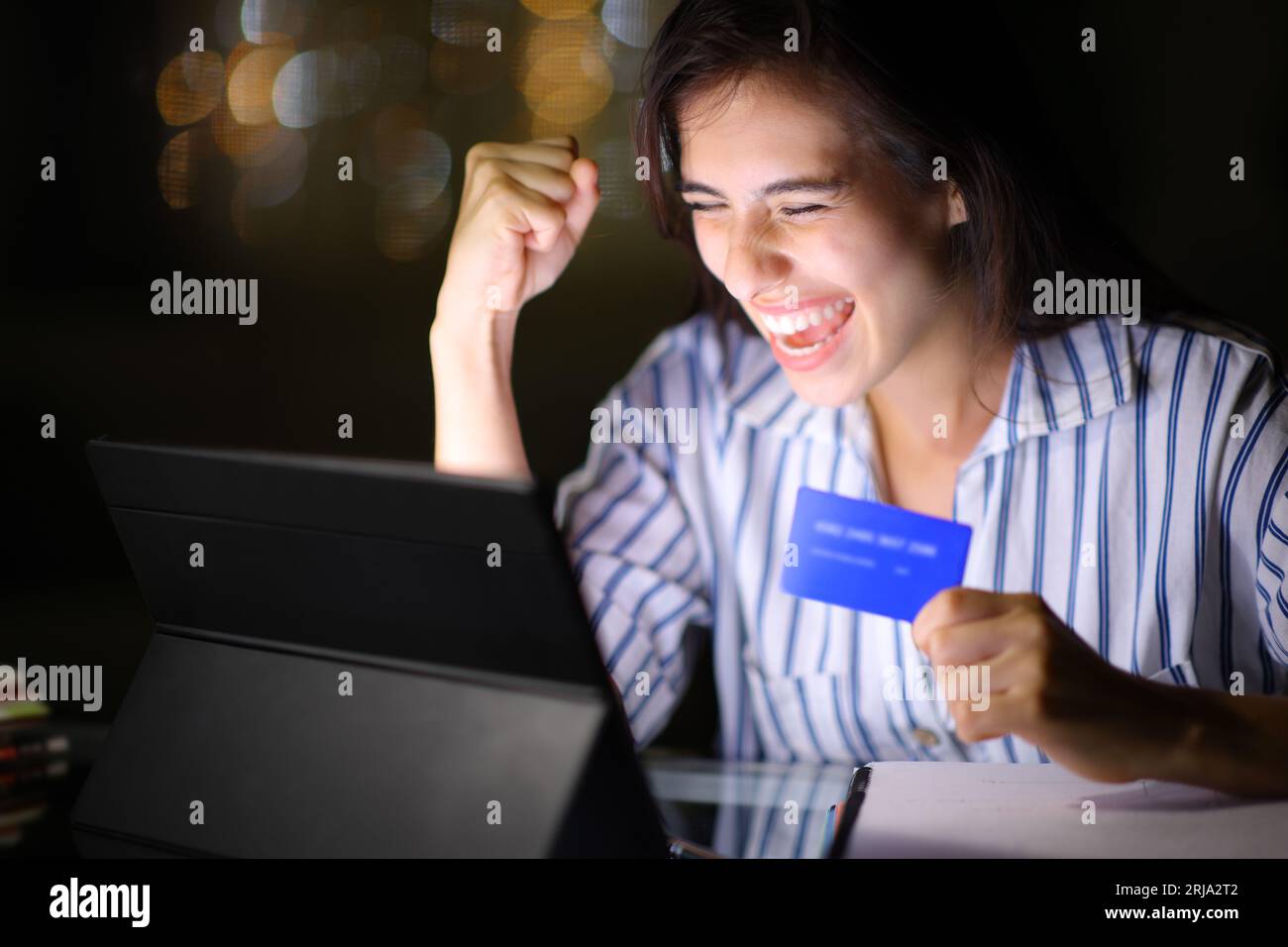 Mujer emocionada que compra en línea con la tableta y la tarjeta de crédito en la noche en casa Foto de stock