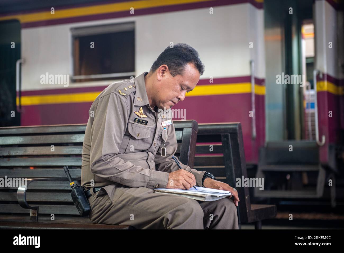 Bangkok, Tailandia - 6 de marzo 2020: Oficial de policía con un uniforme marrón sentado en un banco en la plataforma de la estación de tren de Bangkok, escribiendo diligentemente una nota Foto de stock