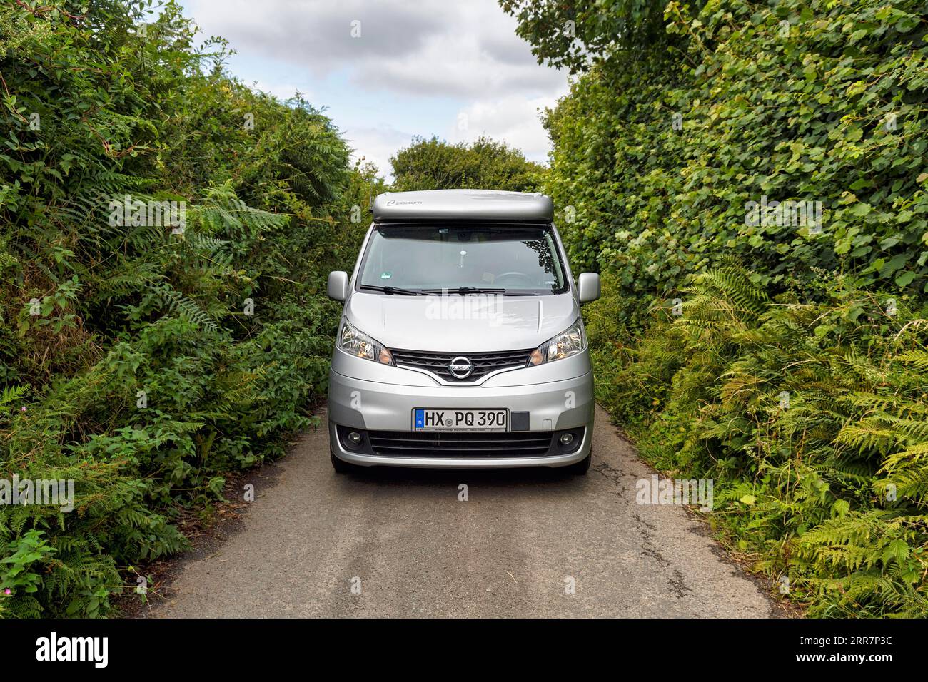 Campervan, Minicamper Nissan NV200 Conduciendo en una carretera rural estrecha de un solo carril, setos altos densos en el borde de la carretera, poca visibilidad, Cornwall, Inglaterra Foto de stock