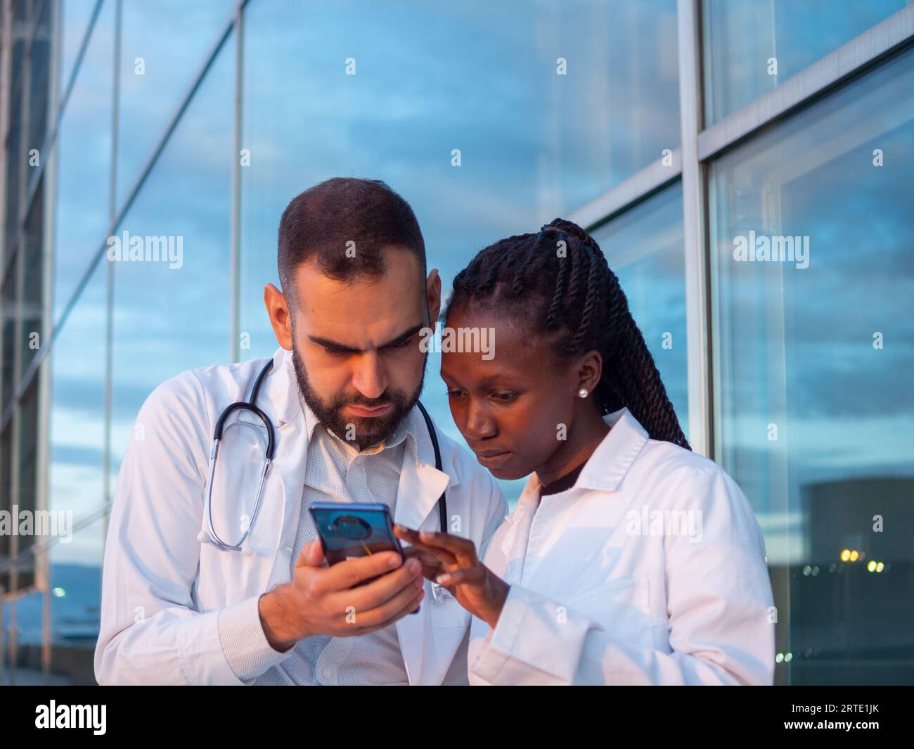 Dos médicos del hospital, una mujer y un hombre, tomando un descanso al aire libre y viendo algo en su teléfono Foto de stock
