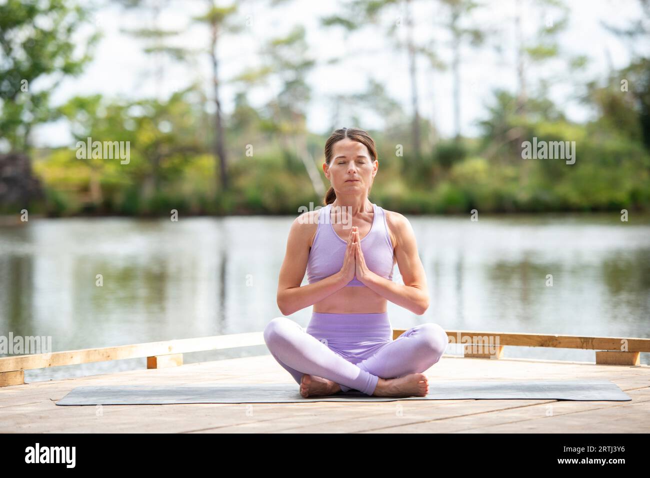 Mujer deportiva practicando yoga en un embarcadero junto a un lago, sentado meditando, parte de la serie. Foto de stock