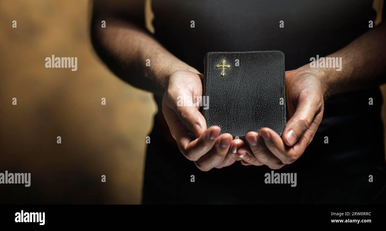 Hombre sosteniendo una vieja pequeña biblia negra en sus manos. Corta profundidad de campo, la nitidez está en la cruz Foto de stock