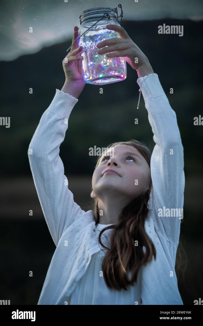 Retrato de una niña mirando hacia arriba en el tarro de cristal con luces de hadas que ella está sosteniendo por encima de su cabeza al atardecer Foto de stock