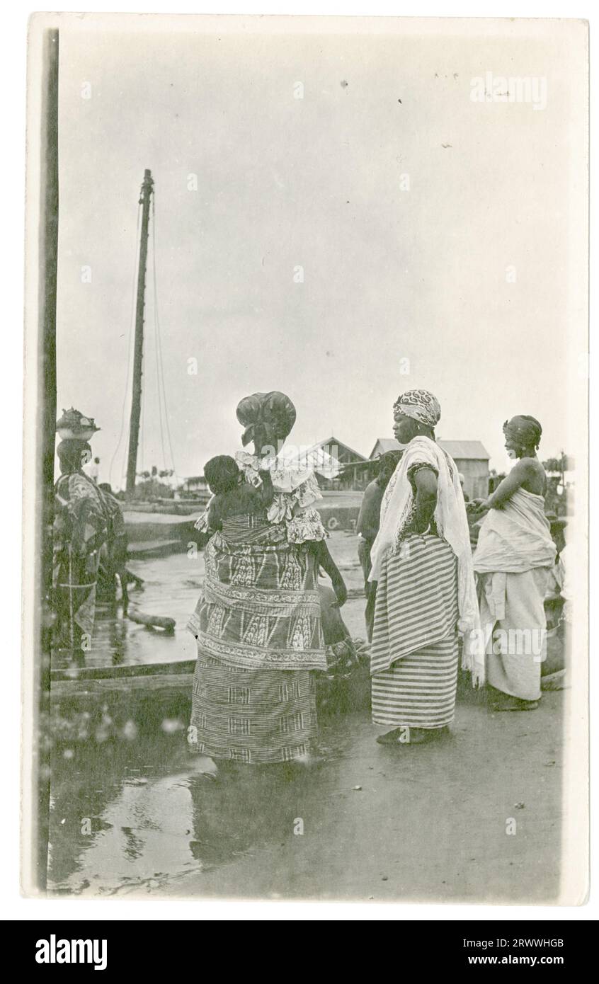 Un pequeño grupo de mujeres africanas se paran junto al puerto, mirando hacia el mar, en un lugar no identificado. Hay edificios junto al muelle y un barco a la vista. Todos están usando vestidos de abrigo impresos y cubiertas de la cabeza y uno tiene un bebé atado a su espalda. Foto de stock