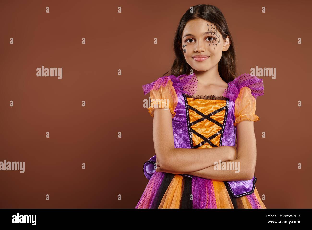 Chica complacida en traje de Halloween con maquillaje de tela de araña de pie con los brazos doblados sobre fondo marrón Foto de stock