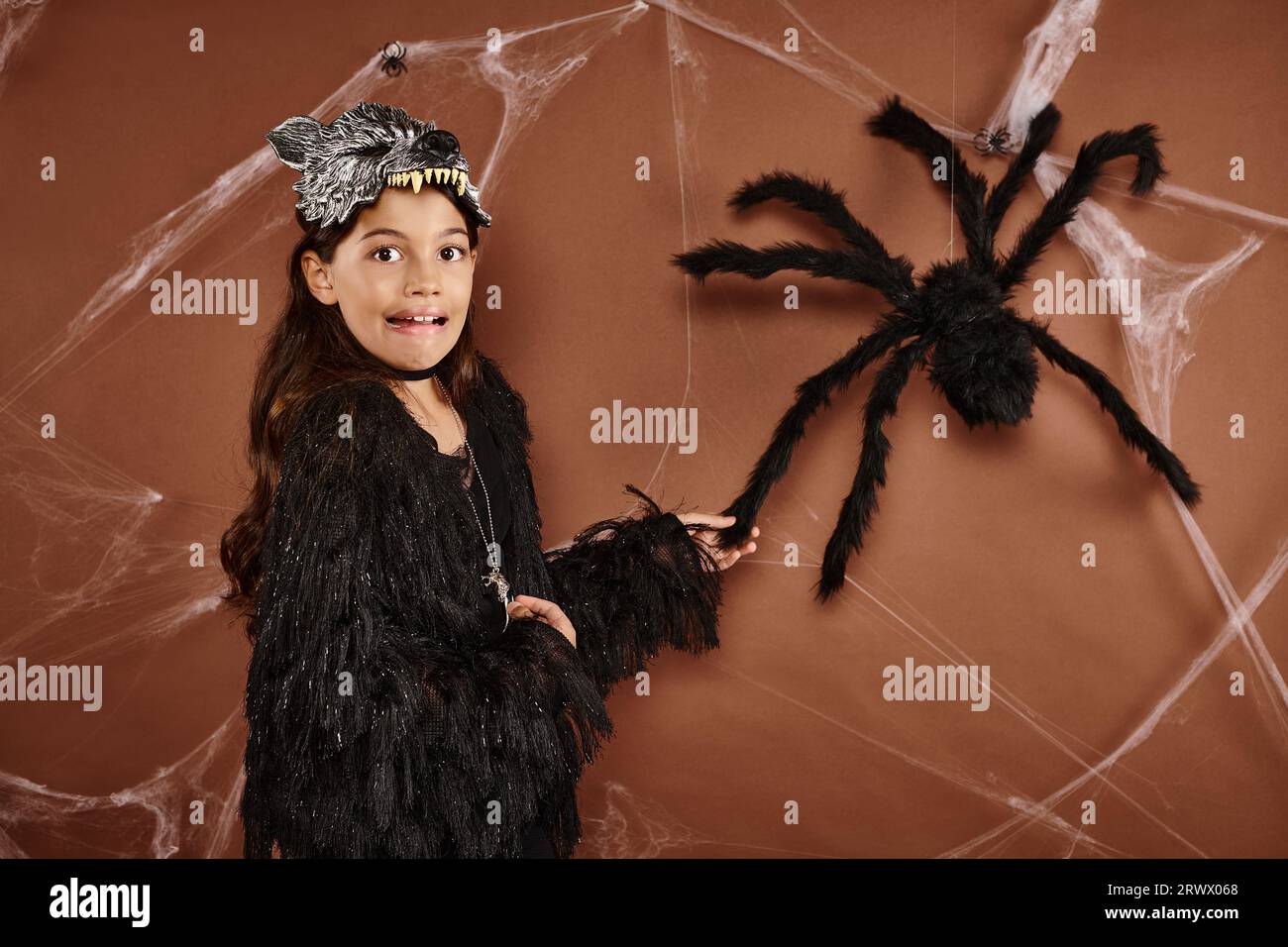Cerrar chica asustada en máscara de lobo y atuendo negro tocando araña en fondo marrón, Halloween Foto de stock
