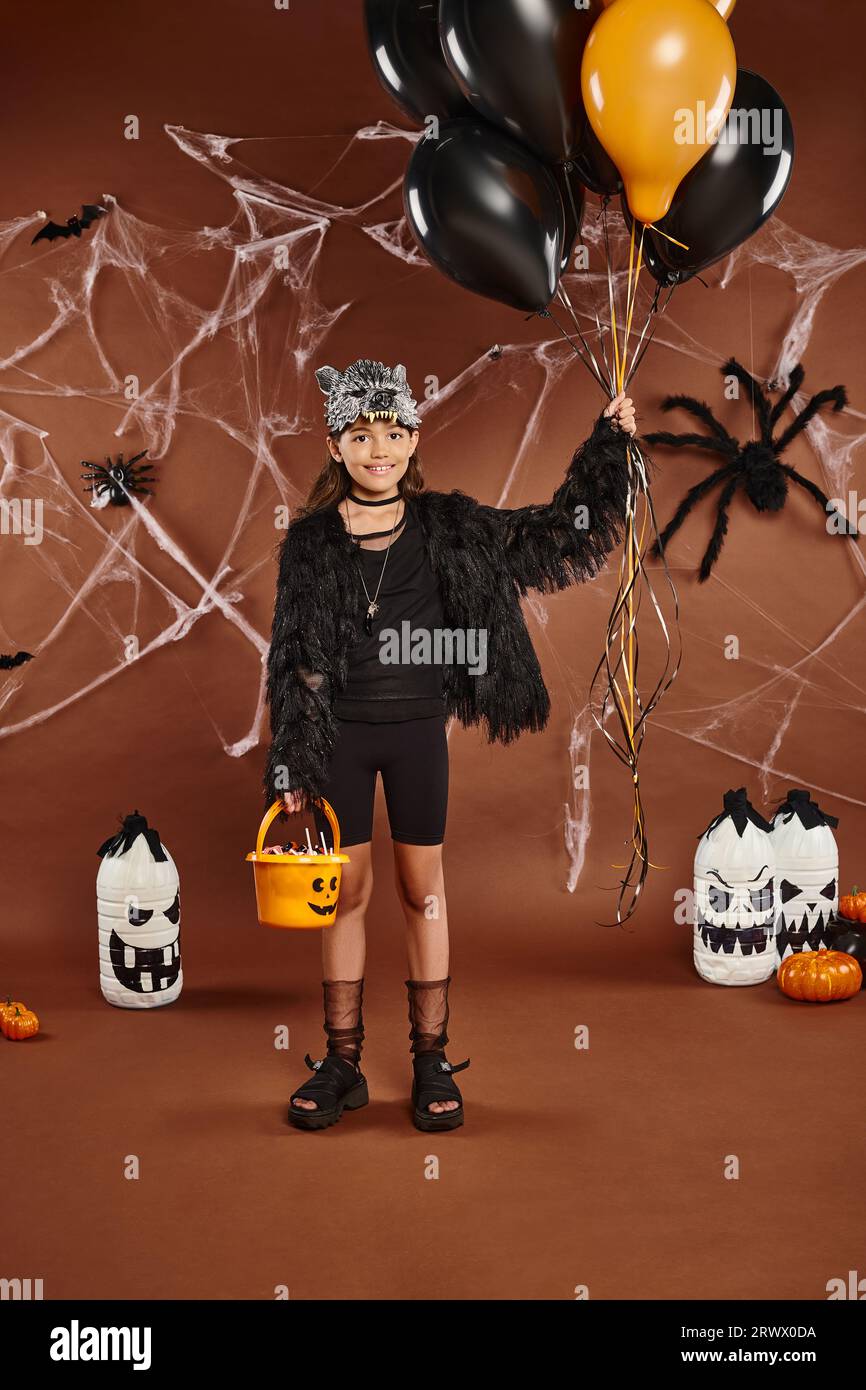 Chica sonriente sostiene globos negros y naranjas y cubo con dulces, concepto de Halloween Foto de stock