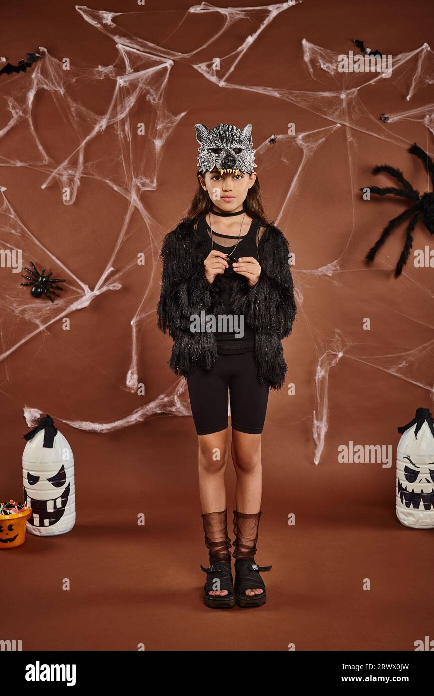Linda chica en traje de piel sintética con máscara de lobo de pie todavía en fondo marrón, concepto de Halloween Foto de stock