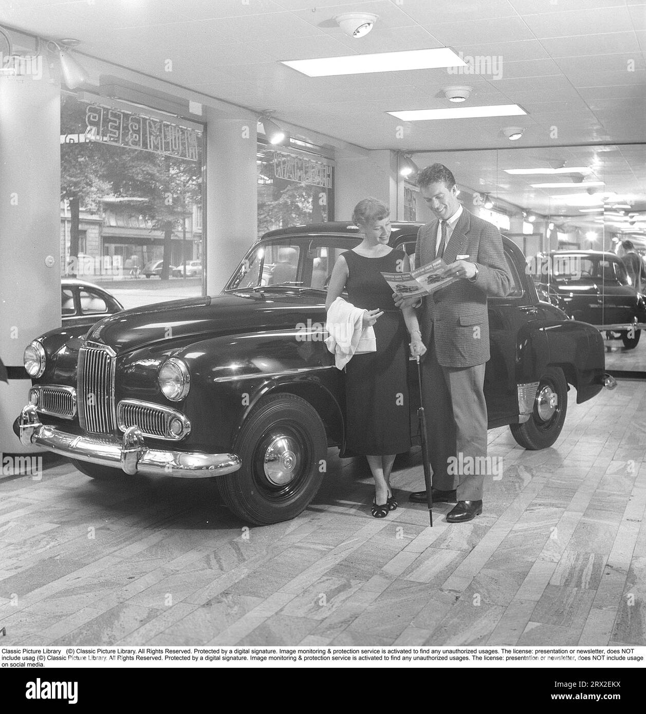 En los años cincuenta. Una joven pareja visita un concesionario de automóviles y el distribuidor que vende la marca de automóviles inglesa Humber. La pareja estudia una carpeta sobre el coche en la habitación donde el coche está estacionado y se ve satisfecho. Están leyendo una carpeta sobre el icónico viaje con el coche entre Londres y Ciudad del Cabo. En 1952 George Hinchcliffe, Arthur Longman y Robbie Walshaw partieron de Londres para conducir a Ciudad del Cabo en un Humber Supersnipe en el tiempo récord de 13 días 9 horas 6 minutos. Suecia 1954. Kristoffersson ref BP39-5 Foto de stock