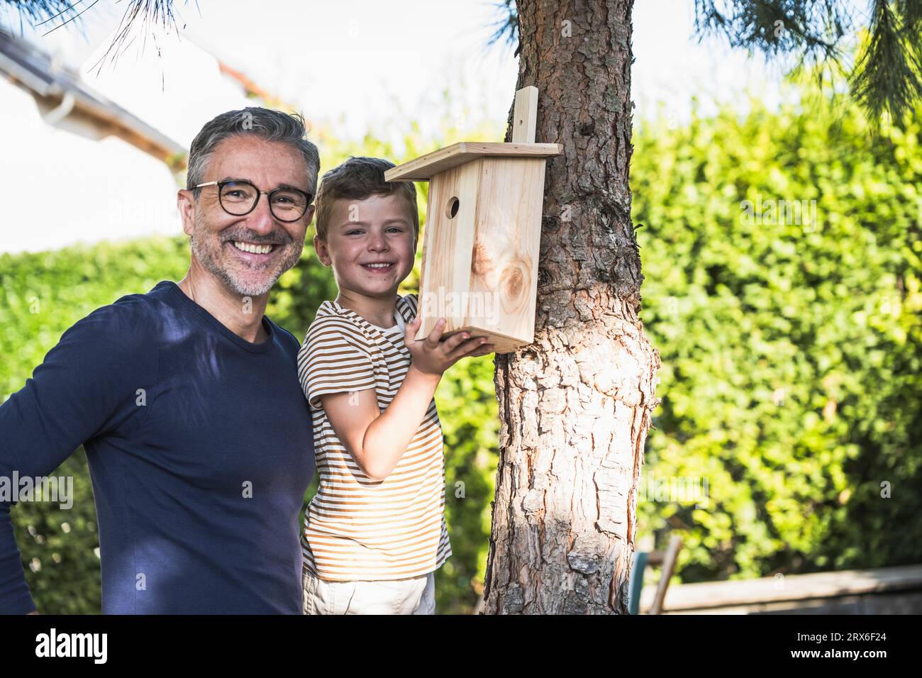 Sonriente hombre y niño de pie junto a la casa de pájaros en el árbol en el patio trasero Foto de stock