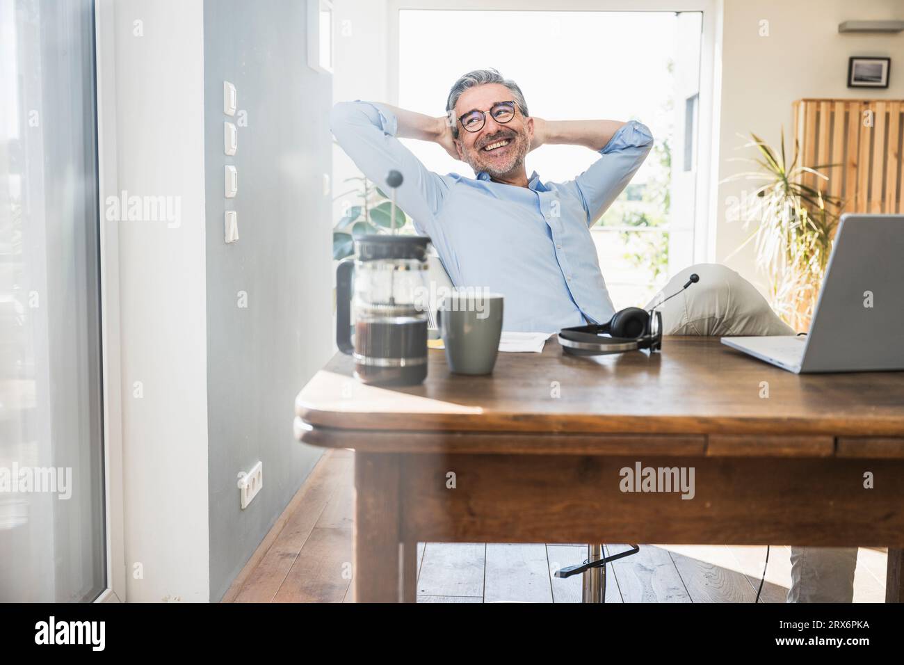 Hombre de negocios feliz con las manos detrás de la cabeza sentado en el escritorio en la oficina en casa Foto de stock