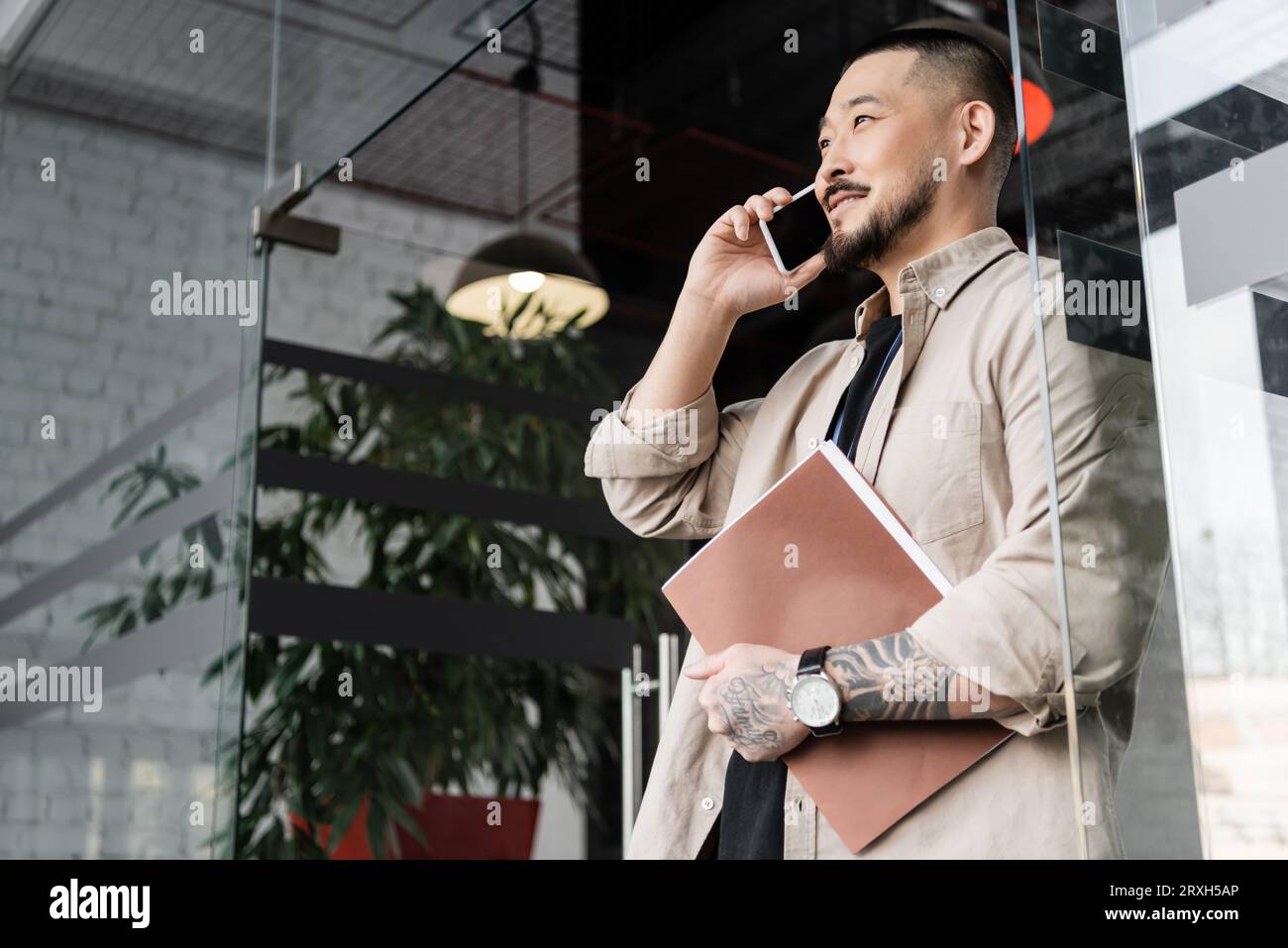 hombre de negocios asiático feliz con carpeta de tatuaje que sostiene y habla en el teléfono inteligente cerca de la puerta de vidrio Foto de stock
