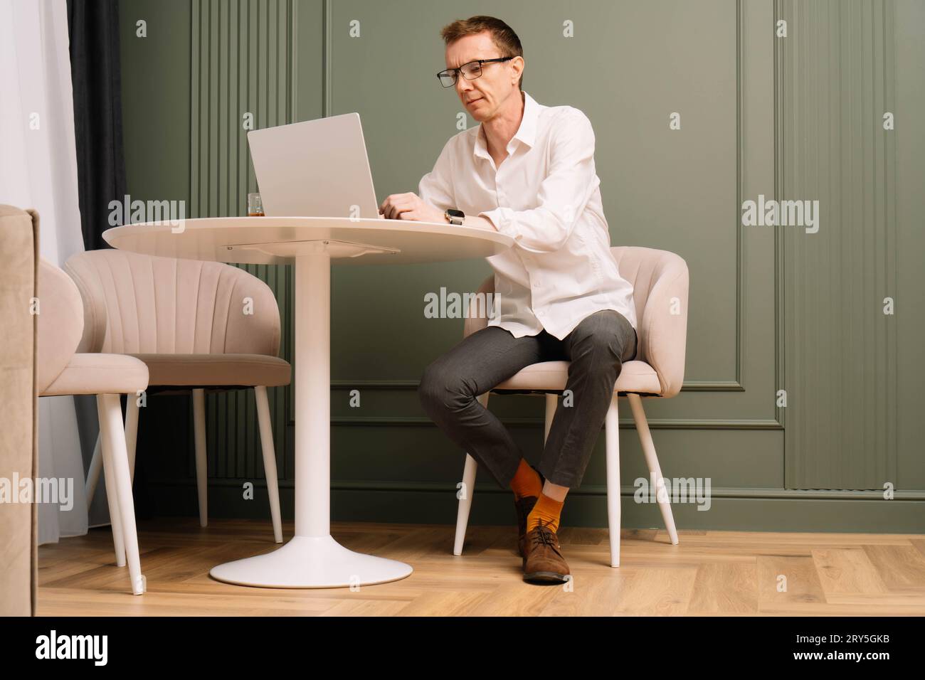 Pensativo hombre de mediana edad en gafas y camisa blanca sentado en la mesa y trabajando en el ordenador portátil. Hombre de negocios industrioso en calcetines naranjas con entusiasmo Foto de stock