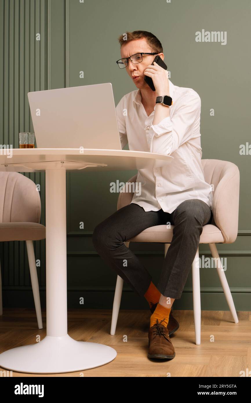 Hombre de negocios elegante sentado en la mesa y hablando por teléfono inteligente mientras funciona en la computadora. Hombre maduro en gafas, camisa blanca y calcetines naranjas de trabajo Foto de stock