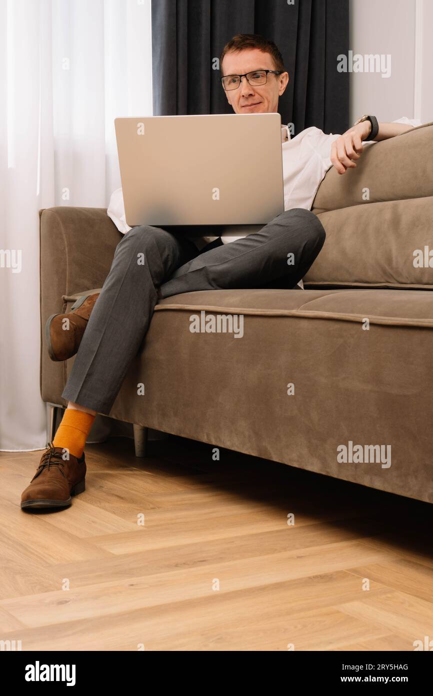 Hombre de negocios de mediana edad en gafas sentado cómodamente en el sofá y trabajando en la computadora. Hombre en camisa blanca, pantalón gris oscuro y calcetines naranja look Foto de stock
