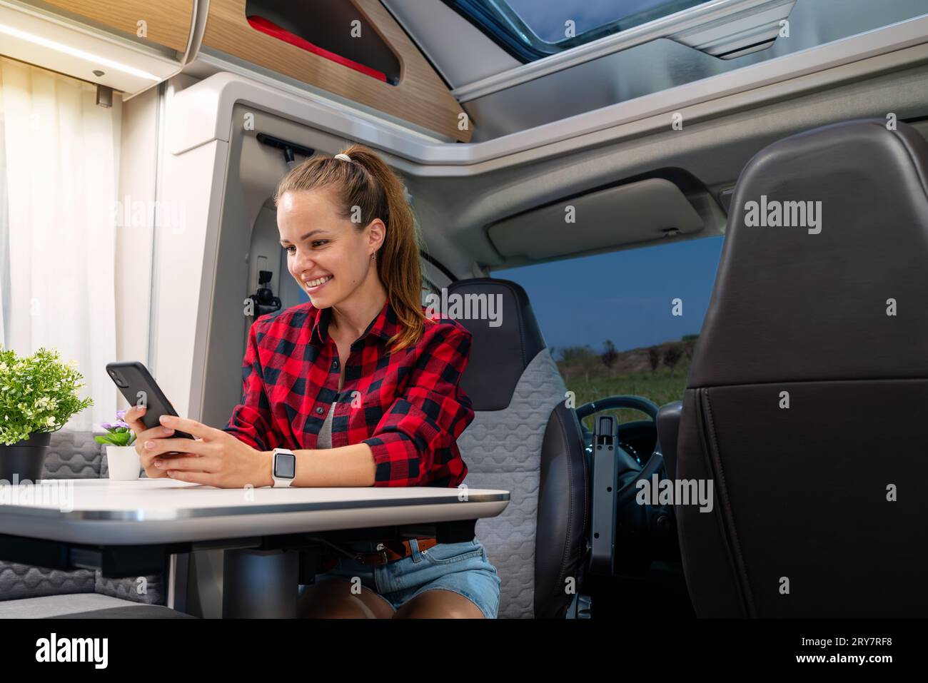 Mujer morena feliz sentada en la mesa en caravana y usando su teléfono inteligente. Alquiler de RV. Autocaravana. Viajar con remolque para autocaravanas. Foto de stock