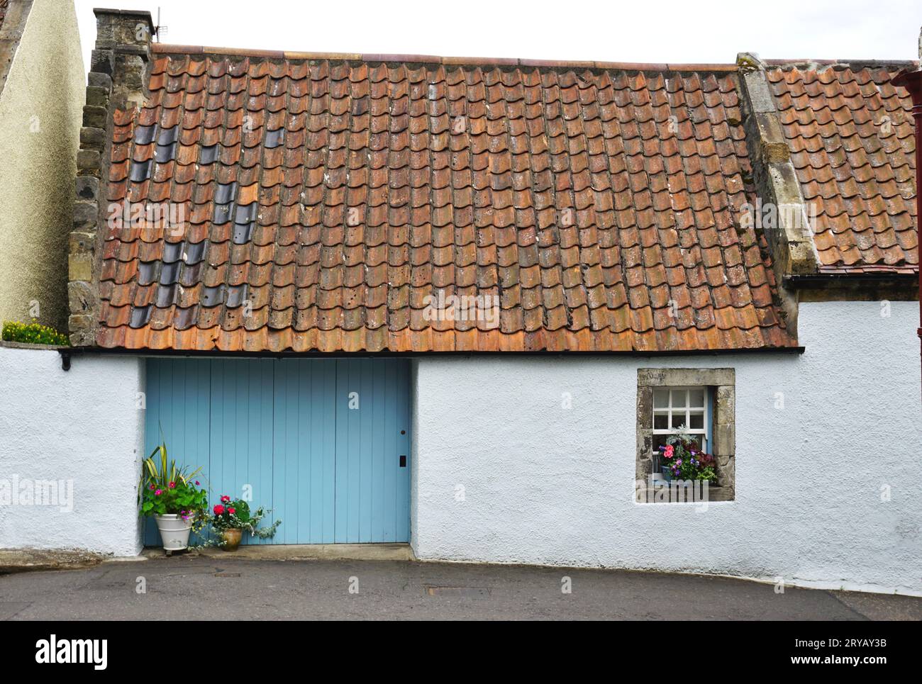 Pintoresca casa de campo con paredes de estuco encaladas, flores en macetas en el alféizar de la ventana y en frente de un panel de puerta azul pálido y techo de baldosas de arcilla vintage Foto de stock