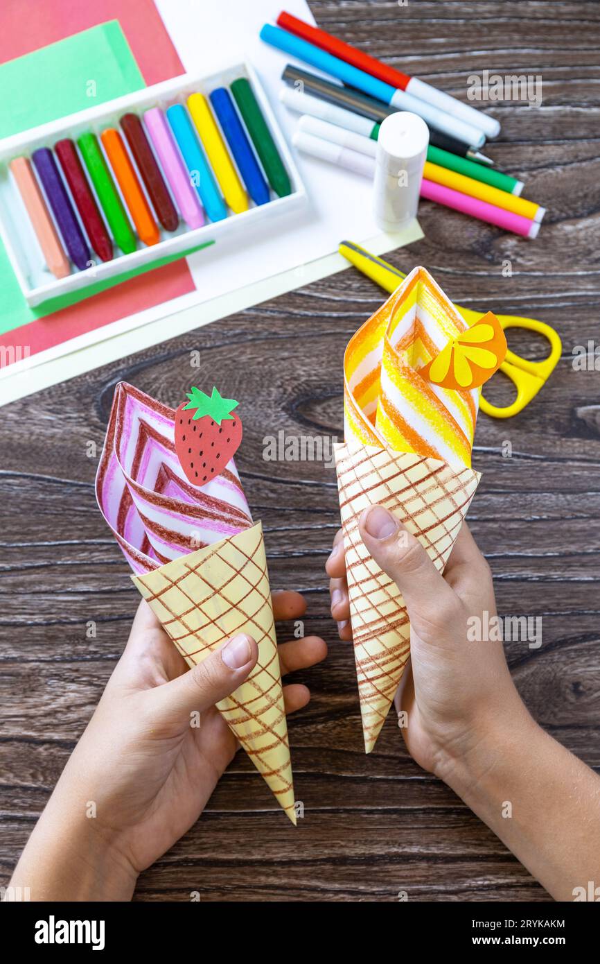 En las manos de un niño Paper Fan helado de papel en una mesa de madera. Proyecto de verano de arte infantil, hecho a mano, manualidades para niños Foto de stock