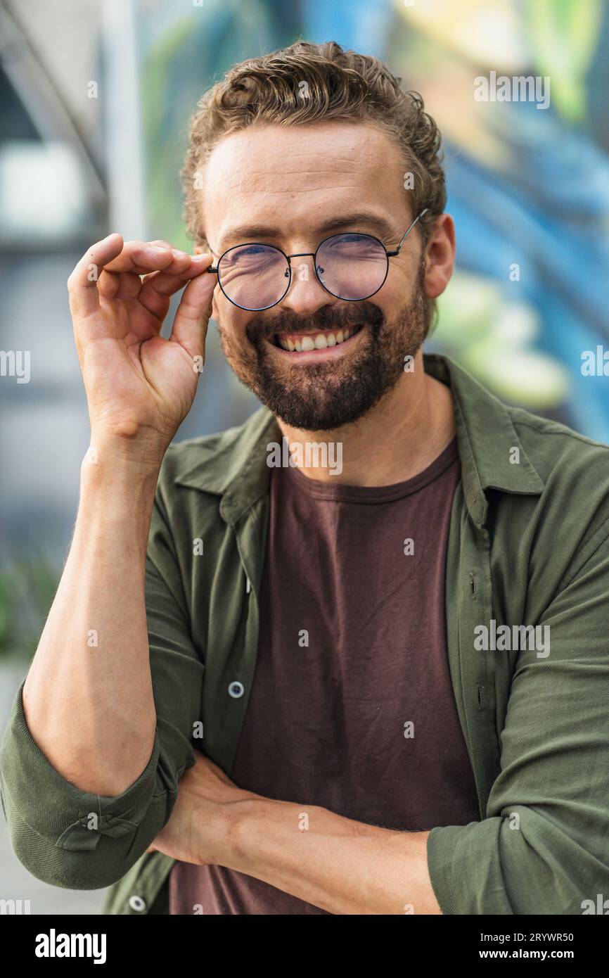 Hombre feliz con sonrisa cálida, sosteniendo gafas. La expresión alegre irradia positividad y satisfacción, mostrando el sentido del sentado interior Foto de stock