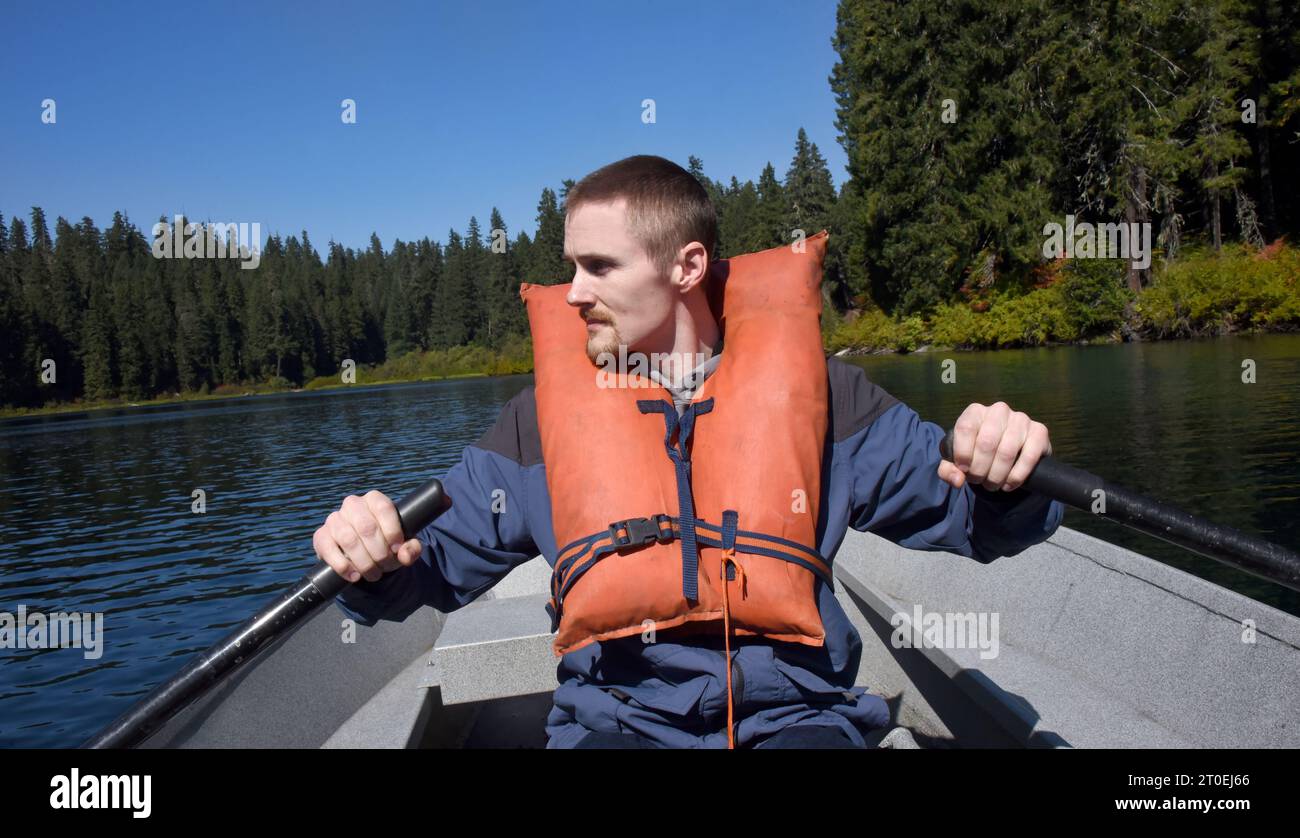 Joven atractivo, con un chaleco salvavidas naranja, rema a través de Clear Lake en Oregon. El cielo es azul. Foto de stock