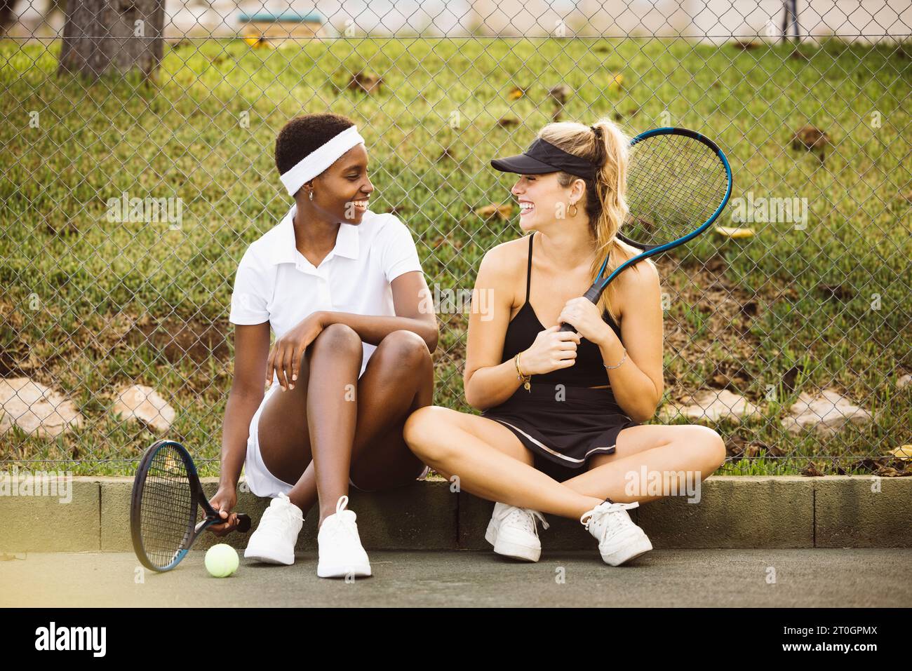 Mujeres hermosas jóvenes con ropa de tenis, sentadas en el suelo de una cancha de tenis y charlando justo antes de comenzar un juego de tenis. Mujeres deportivas gett Foto de stock