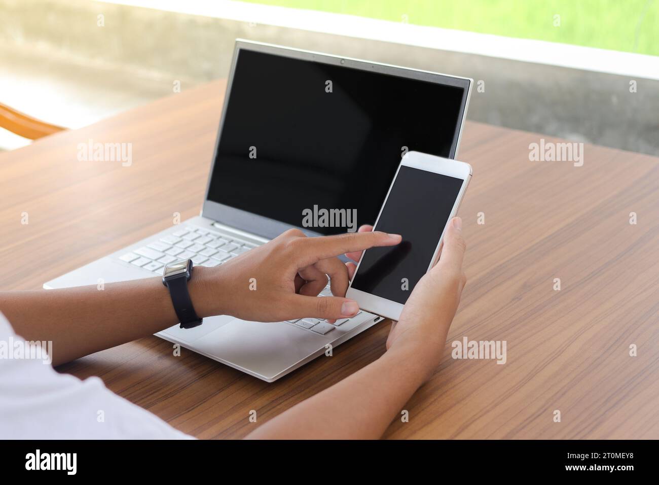 Cierre de la mano del hombre de negocios tocando la pantalla en blanco del teléfono móvil con el ordenador portátil al lado en la mesa. Pantalla de smartphone y portátil simulada Foto de stock