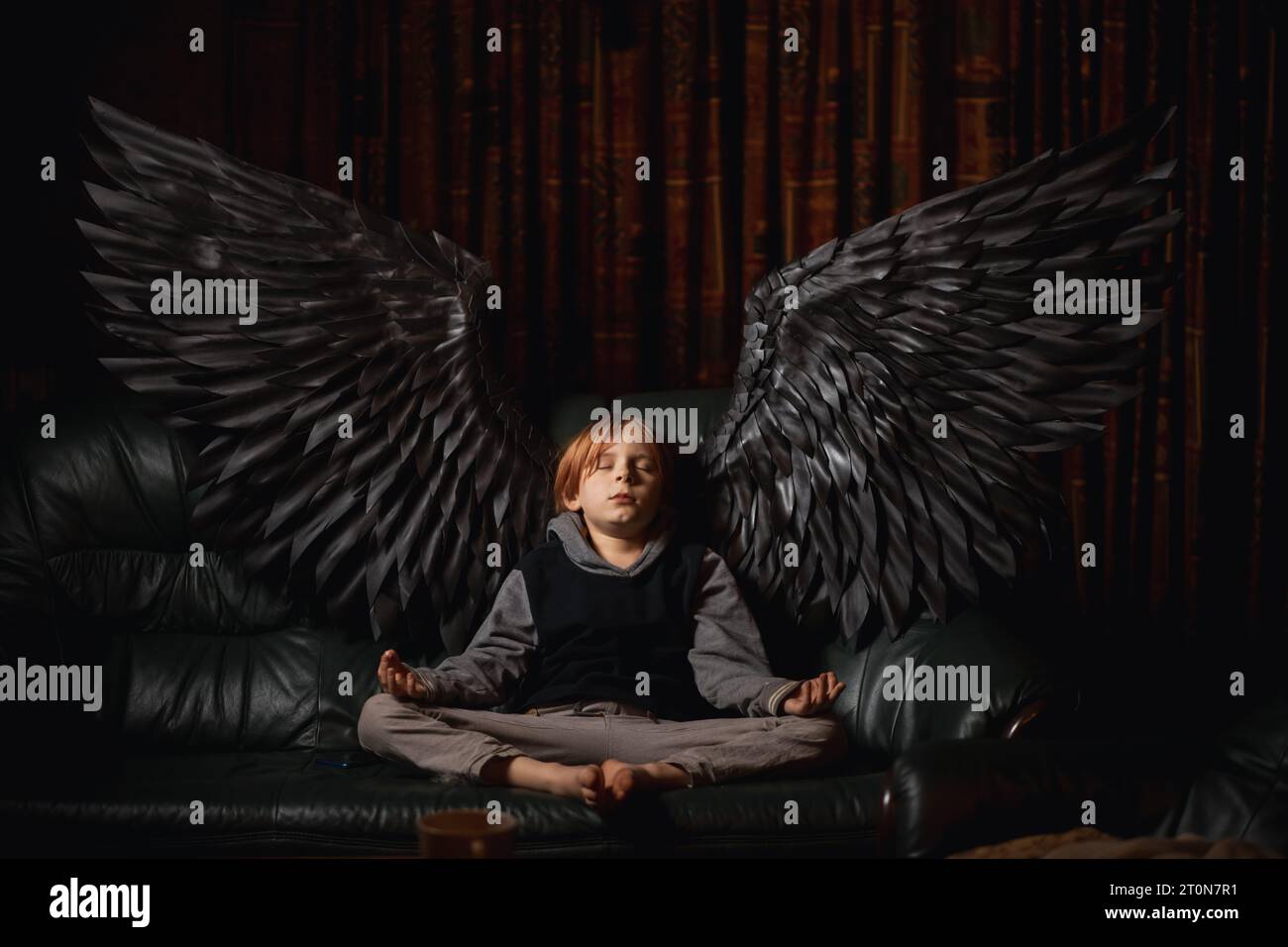 Un joven, de 9-10 años, se sienta en un sofá en una habitación oscura, meditando con sus alas de ángel negras extendidas. El chico se ve pacífico y sereno, como Foto de stock