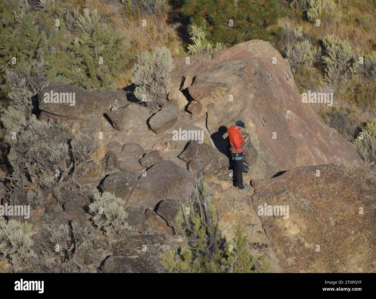 Escalador de montaña se encuentra en el acantilado en Smith Rock State Park, en Oregón, y prepara su equipo para repeler. Lleva puesta una sudadera naranja. Foto de stock