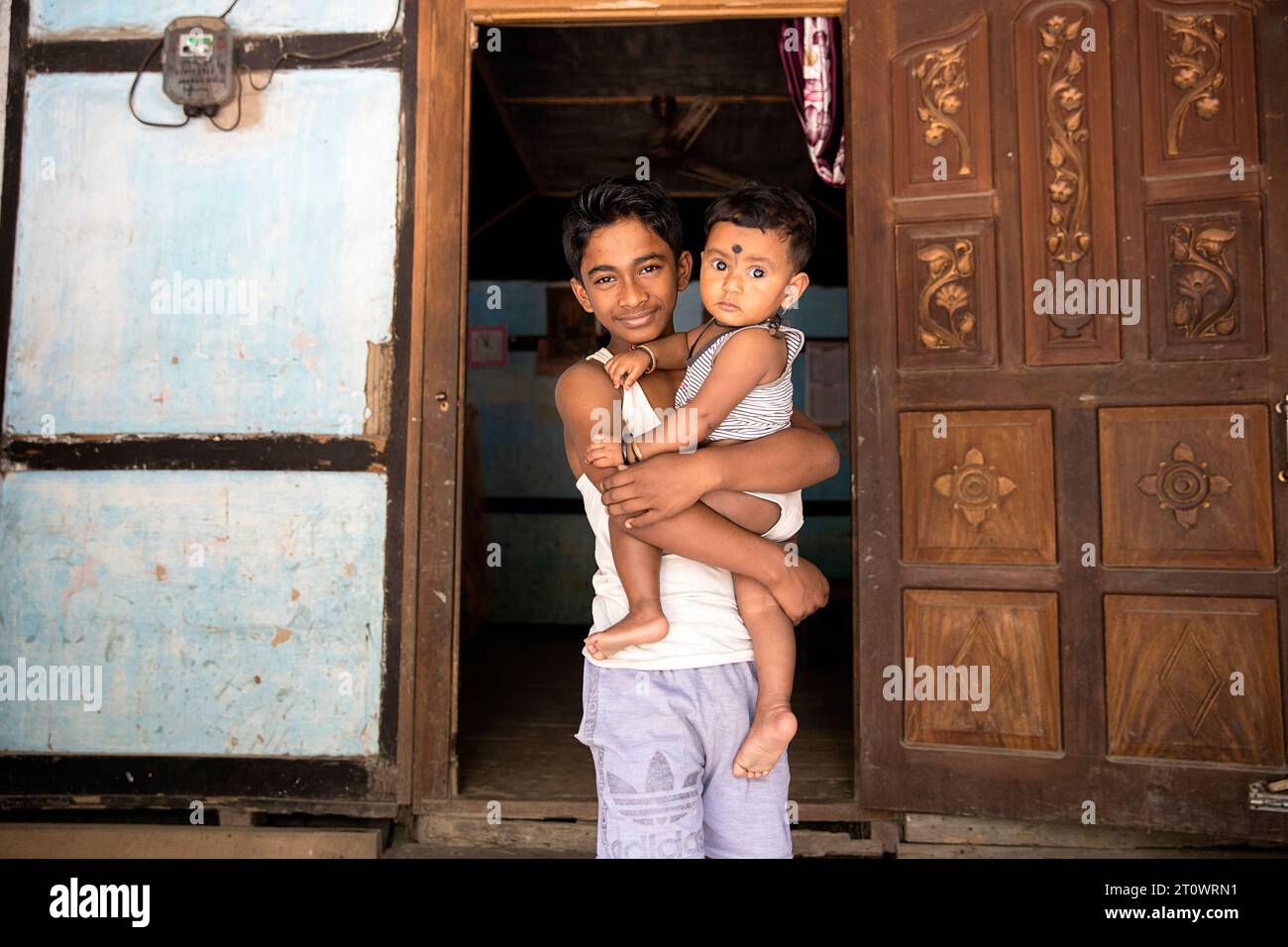 Niño de pie con su hermano pequeño en sus manos en su casa en un pequeño pueblo en la isla de Majuli, Assam, India Foto de stock