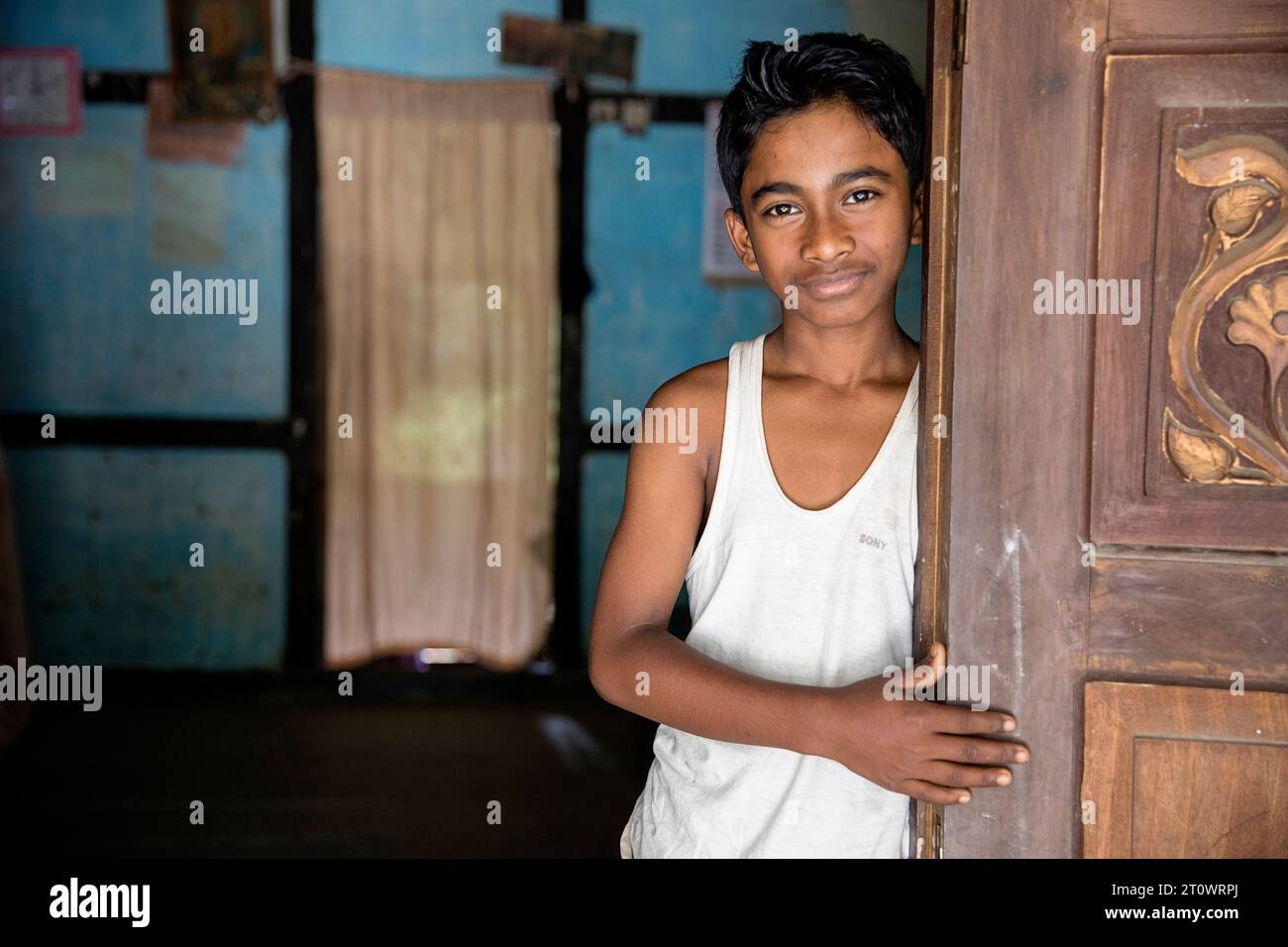 Niño de pie cerca de la puerta de madera tradicional en su casa en un pequeño pueblo en la isla de Majuli cerca del río Brahmaputra en Assam, India Foto de stock