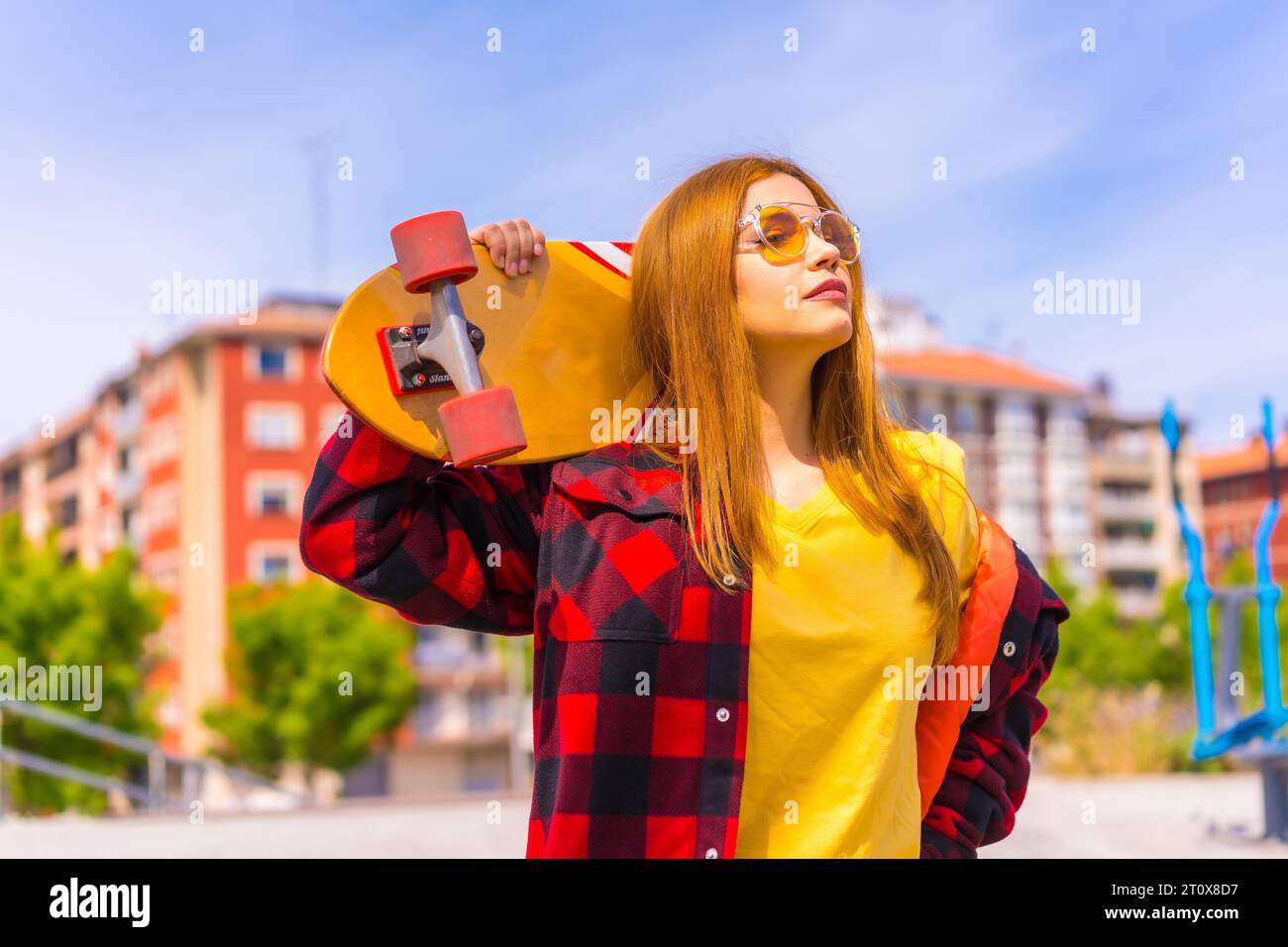Mujer skater en una camiseta amarilla, camisa de cuadros rojos y gafas de sol, pensativamente mirando a la derecha Foto de stock