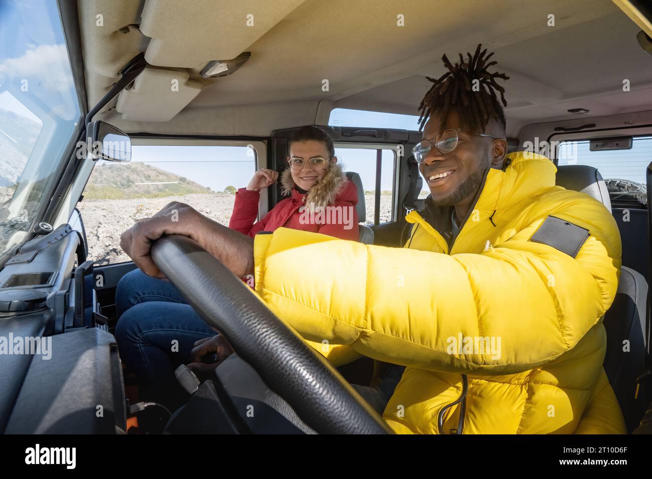 Un hombre africano con chaqueta de excursionista conduce un vehículo todoterreno, con su compañera caucásica mirándolo con una cálida sonrisa. Foto de stock