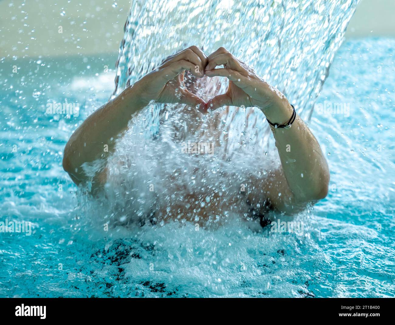 Mujer detrás de una cascada en una piscina DE SPA y haciendo una forma de corazón con sus manos mojadas en Suiza Foto de stock