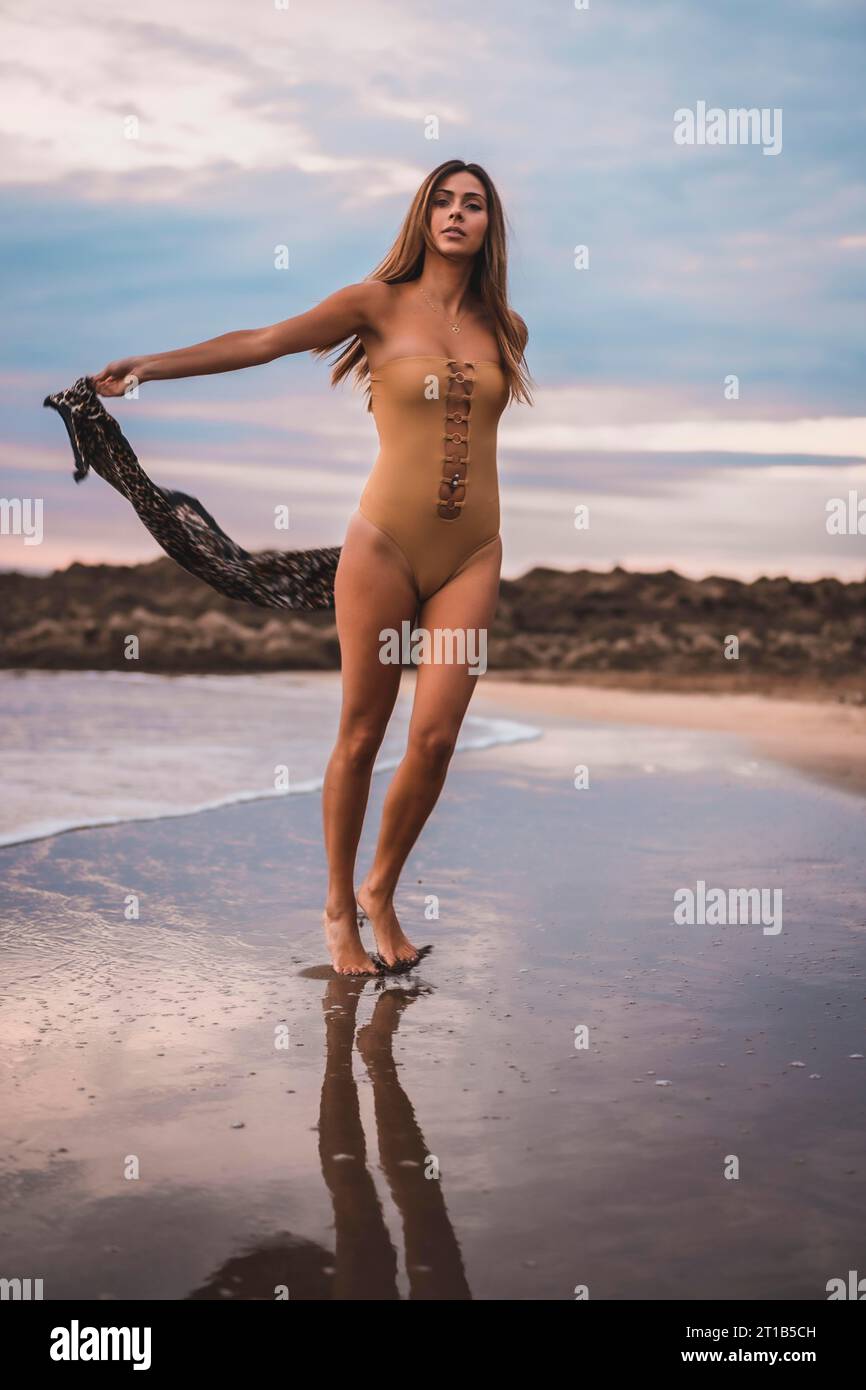 Una mujer morena con un traje de baño y un pareo caminando por la playa en verano en baja mar Foto de stock