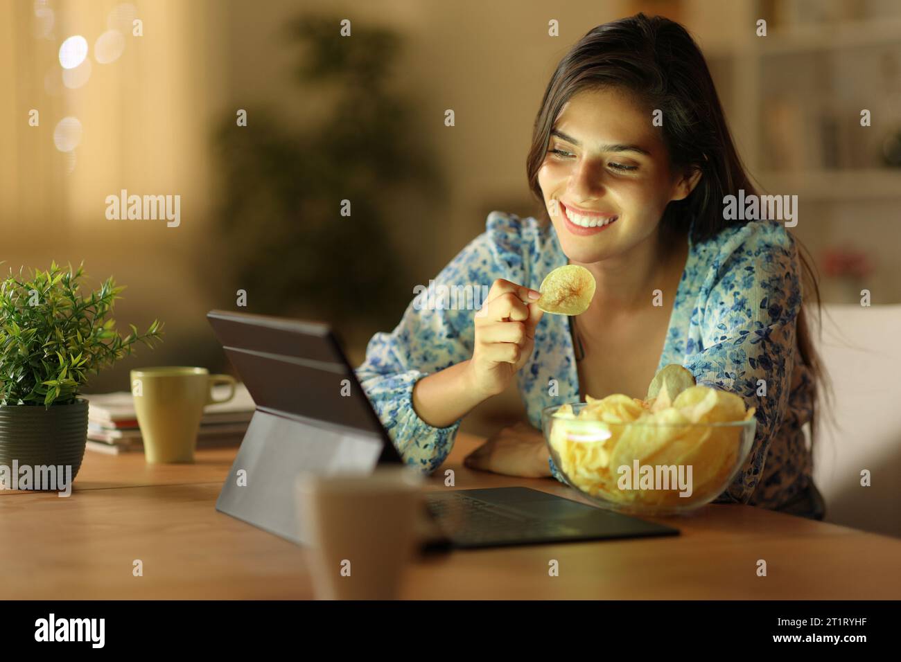 Mujer feliz en la noche viendo contenido en línea comiendo patatas fritas en casa Foto de stock