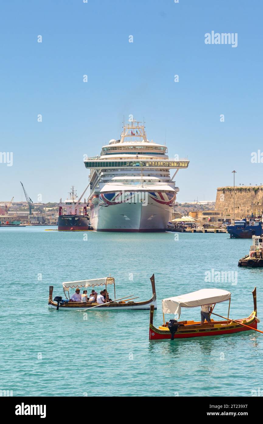 Valletta, Malta - 3 de agosto de 2023: Pequeños barcos de madera que llevan a los turistas a través del puerto en la ciudad de La Valeta. En el fondo es un crucero Foto de stock