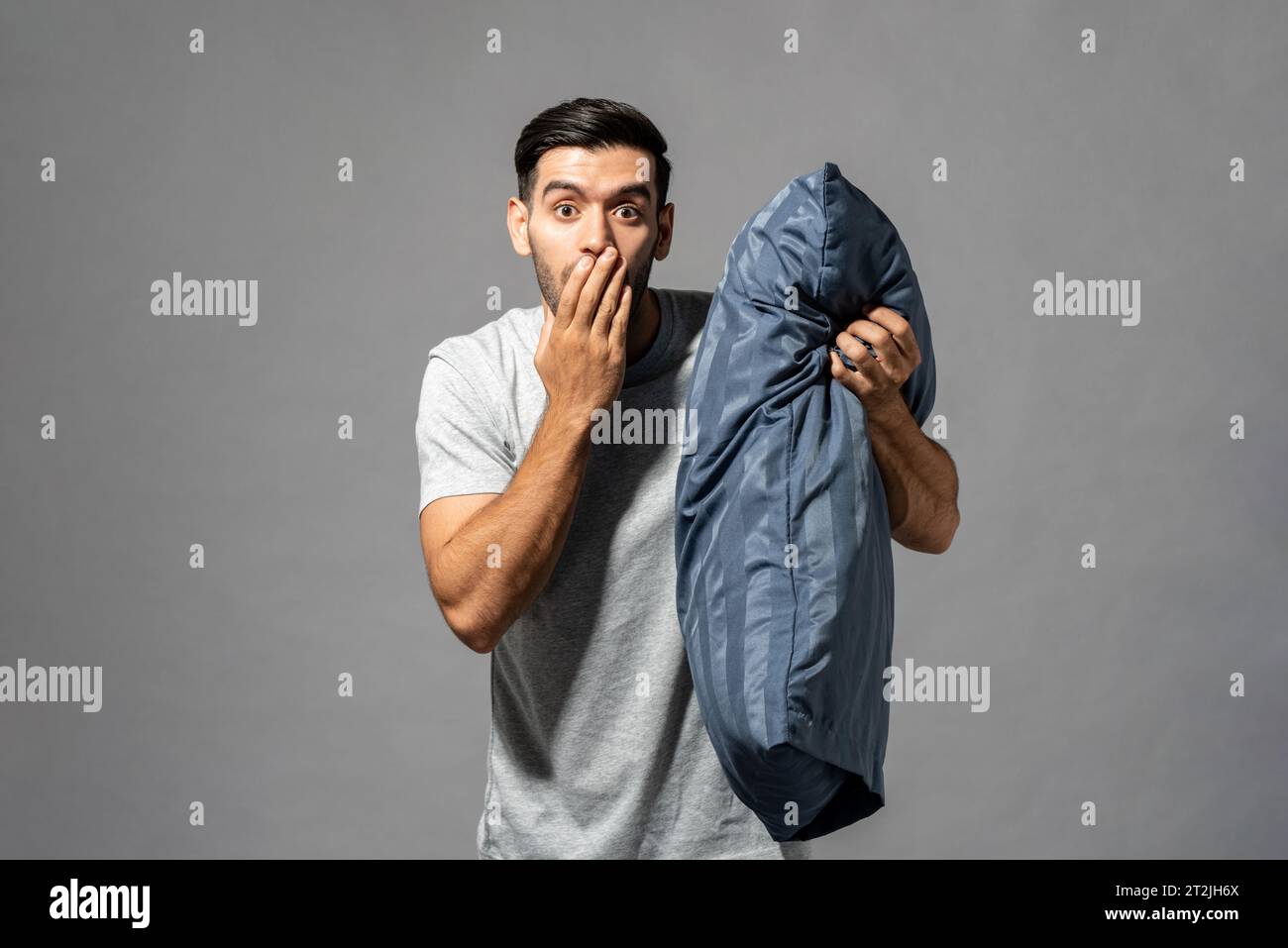 Hombre caucásico asombrado con la almohada suave que cubre la boca y que mira a la cámara sobre fondo gris en estudio ligero Foto de stock