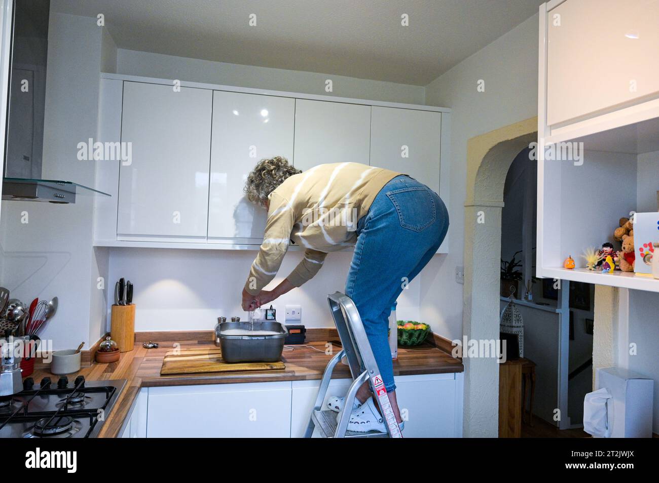 Mujer de mediana edad en una escalera limpiando sus armarios de cocina con tazón de agua jabonosa Foto de stock