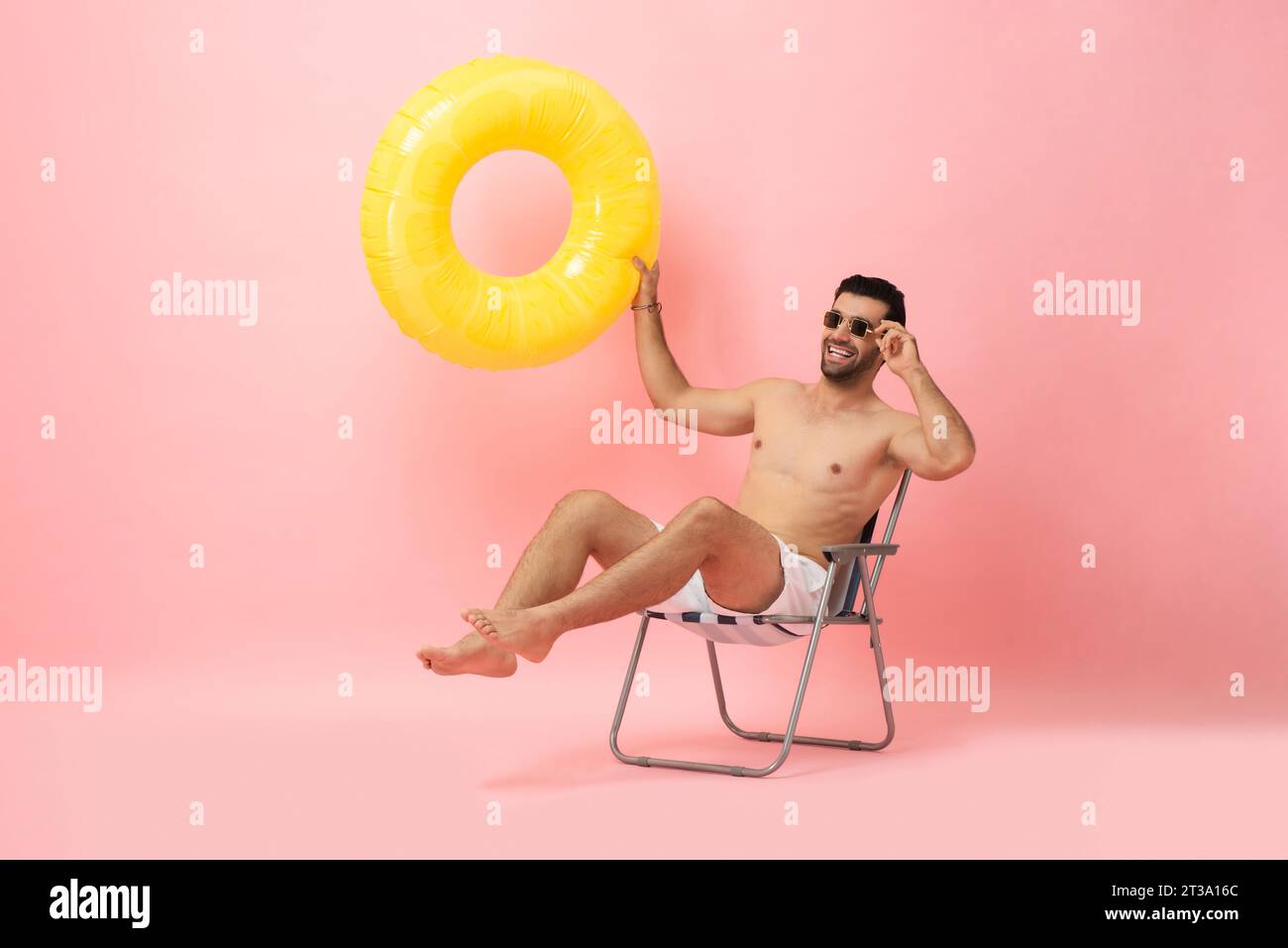 Hombre turístico caucásico sin camisa feliz que se sienta en la silla de la playa que sostiene el anillo de la natación en fondo aislado del tiro del estudio del color rosado, concepto de la vocación del verano Foto de stock