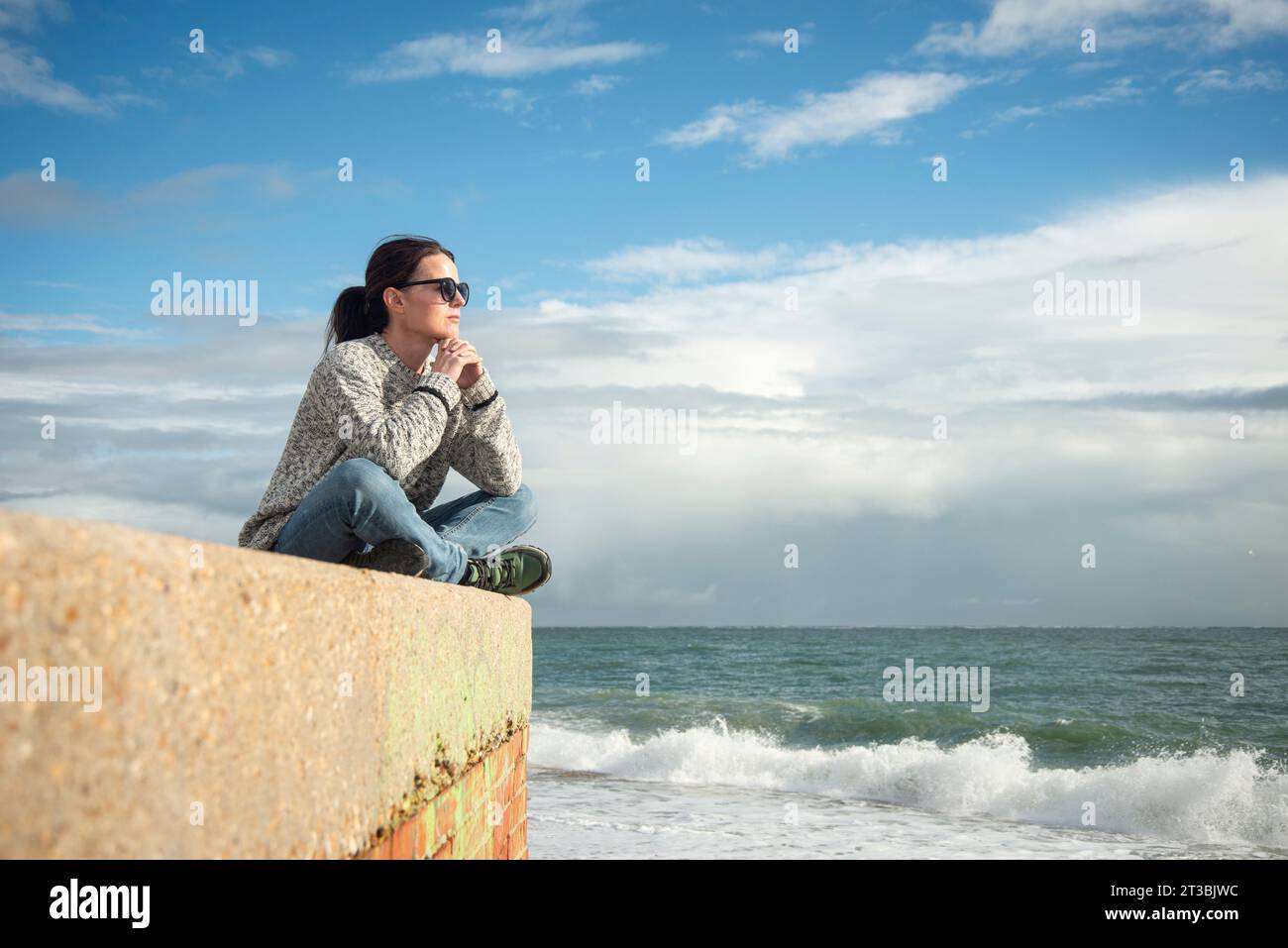 mujer que lleva un suéter y gafas de sol que se relaja y disfruta del invierno, sol del otoño junto al mar Foto de stock